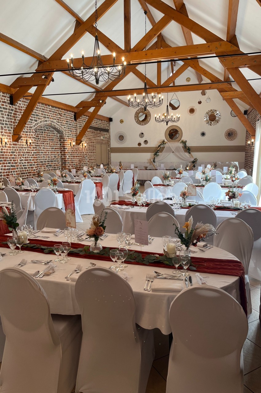 Salle de réception décorée avec des tables dressées, nappes blanches, centres de table floraux et poutres apparentes.