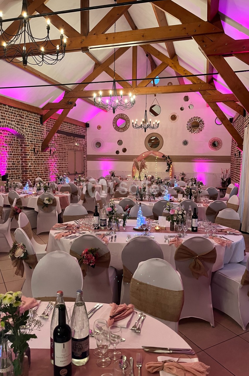 Salle de réception décorée pour un événement, avec tables dressées, nappes blanches et éclairage rose.