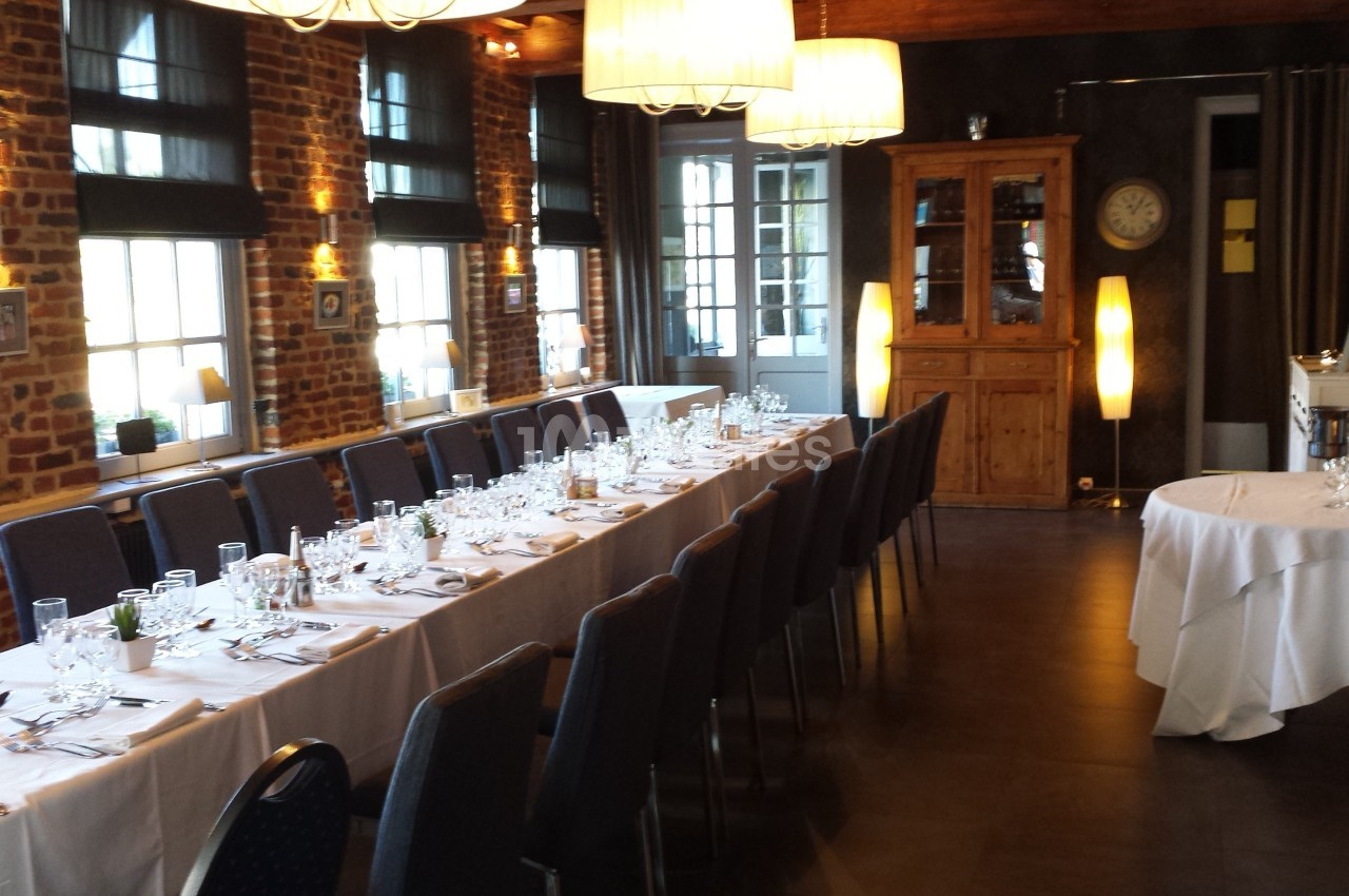 Salle de réception avec longues tables dressées, vaisselle élégante et lumière naturelle entrant par des fenêtres.