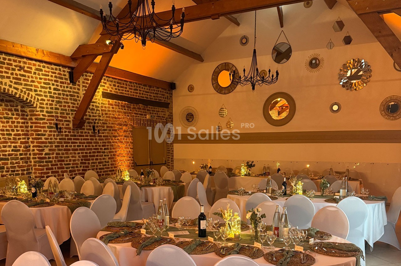 Salle de réception décorée avec des tables dressées, des chaises blanches et des lumières tamisées sous un plafond en…