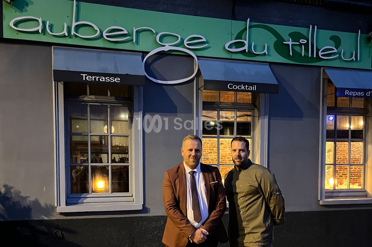 Deux hommes posent devant la façade éclairée d'un restaurant nommé ’Auberge du Tilleul’ en soirée.