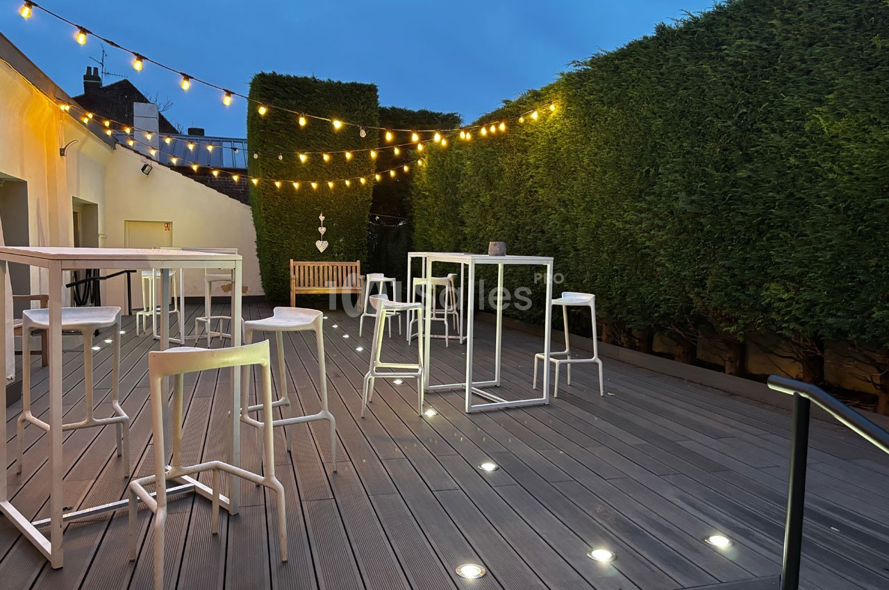 Terrasse en bois avec tables hautes, tabourets blancs, guirlandes lumineuses et haies taillées en soirée.