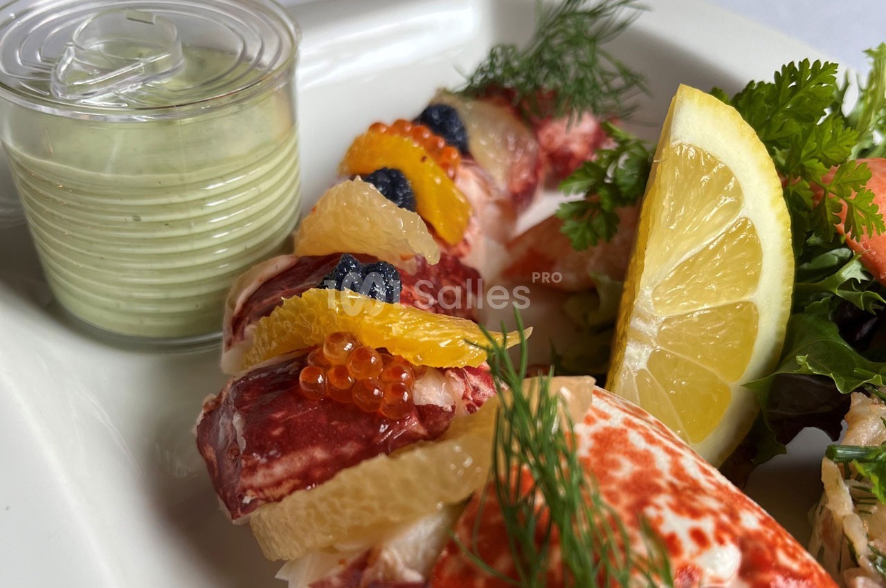 Assiette de homard décorée d'agrumes, œufs de poisson, herbes fraîches, accompagnée d'une sauce verte en verrine.