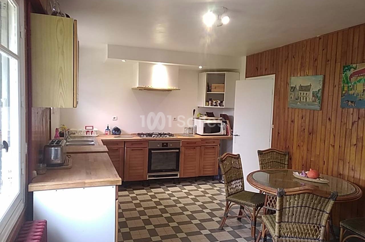 Cuisine avec meubles en bois, table en rotin, sol en carrelage à motifs et mur en lambris, éclairée par une fenêtre.