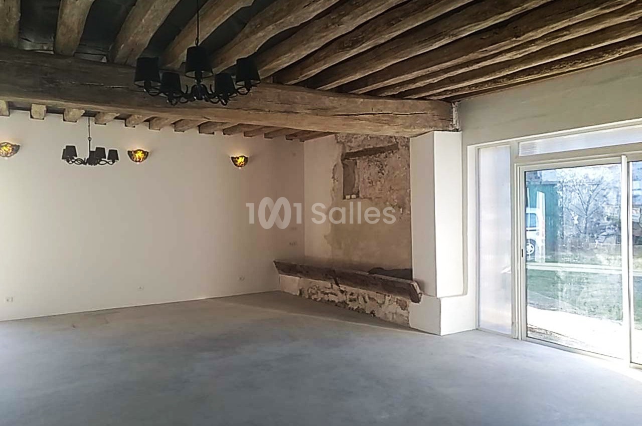 Salle lumineuse avec poutres en bois apparentes, murs clairs, sol en béton et grande baie vitrée donnant sur l'extérieur.
