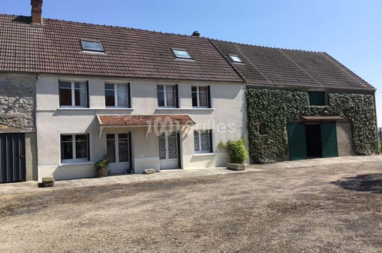 Maison de campagne avec façade claire, toit en tuiles et grange attenante recouverte partiellement de végétation.