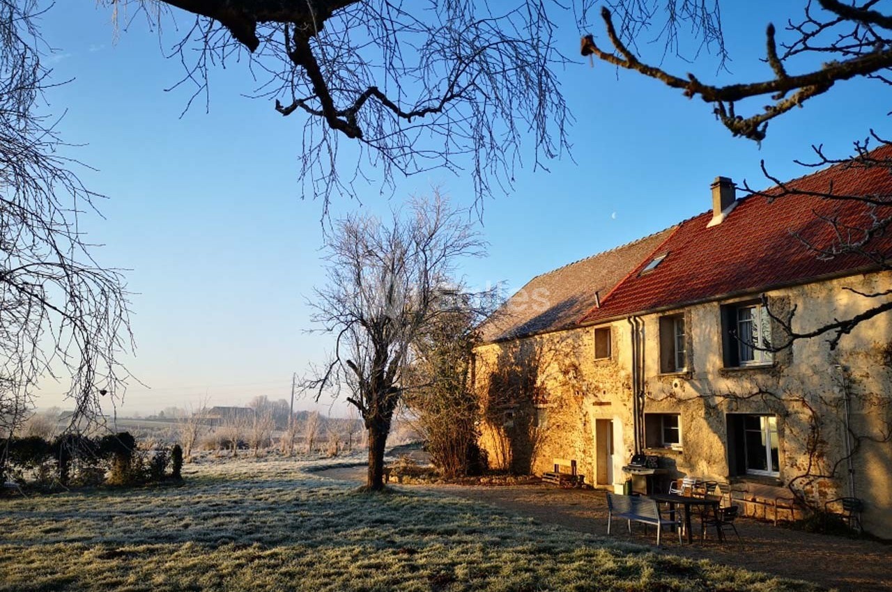 Maison en pierre avec toit rouge entourée d'un jardin givré au lever du soleil, branches d'arbres au premier plan.