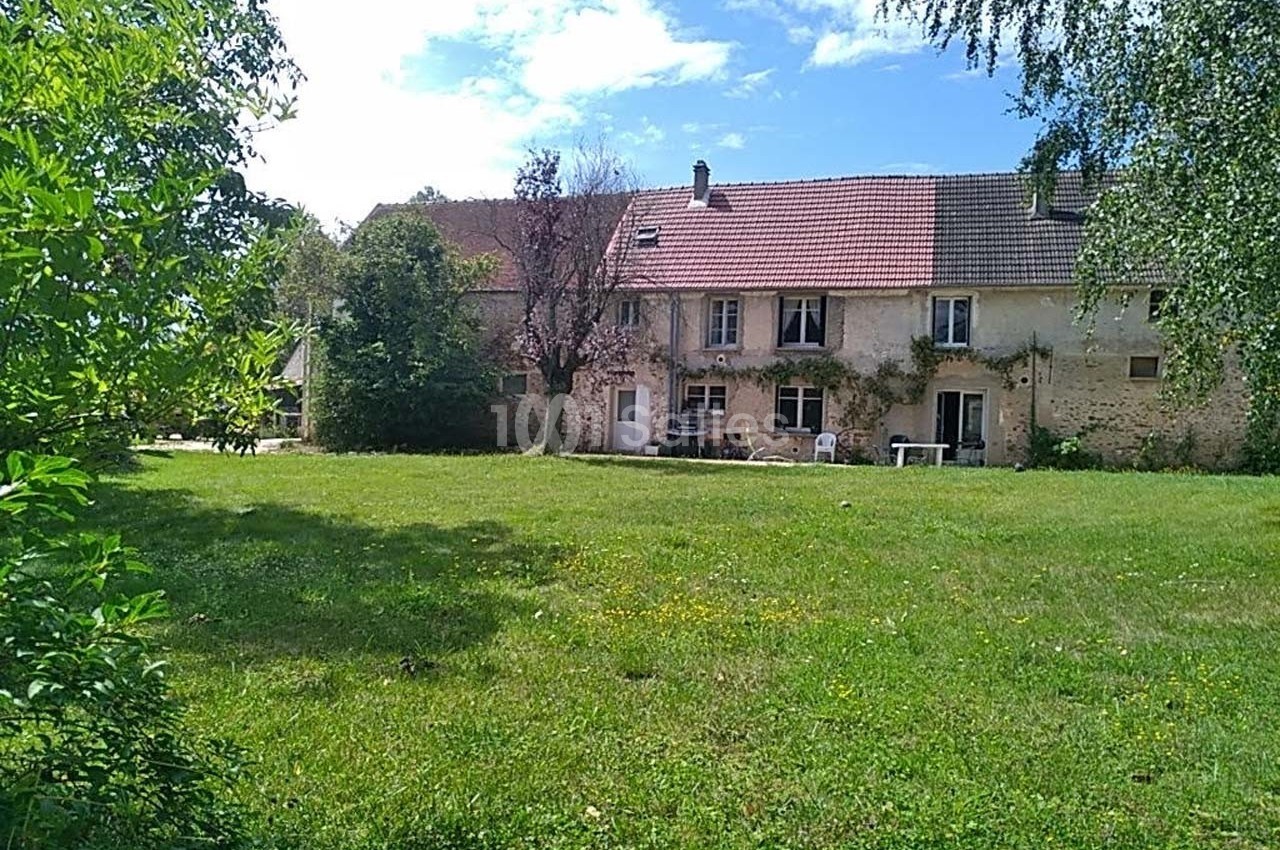 Grande maison en pierre avec toit en tuiles, entourée d'un vaste jardin verdoyant sous un ciel partiellement nuageux.
