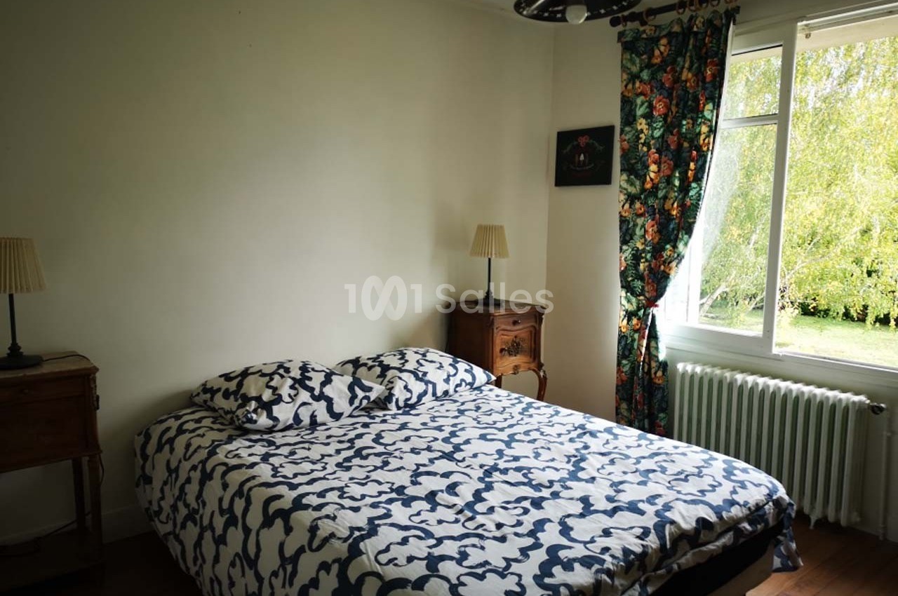 Chambre lumineuse avec un lit double, deux tables de chevet, des rideaux fleuris et une fenêtre donnant sur un jardin.