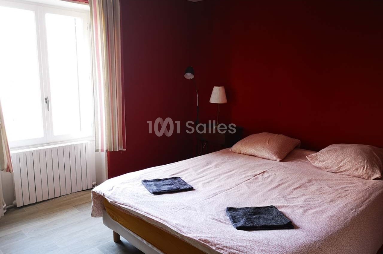 Chambre avec un lit double, draps roses, deux serviettes pliées, mur rouge, fenêtre avec rideaux clairs.