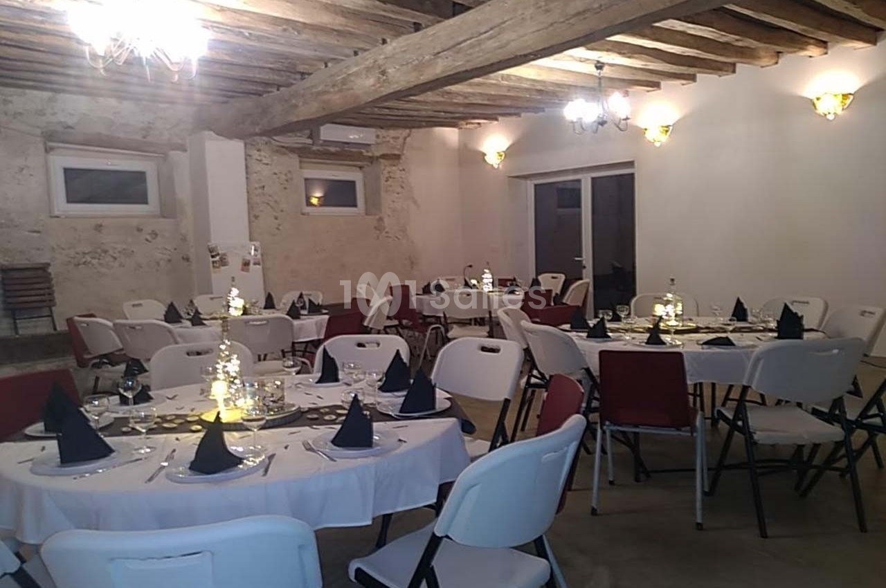 Salle de réception avec tables rondes dressées, nappes blanches, serviettes noires et poutres apparentes au plafond.