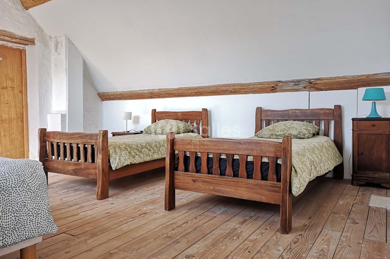 Deux lits en bois avec draps verts dans une chambre mansardée au sol en parquet, éclairée par une lampe de chevet.