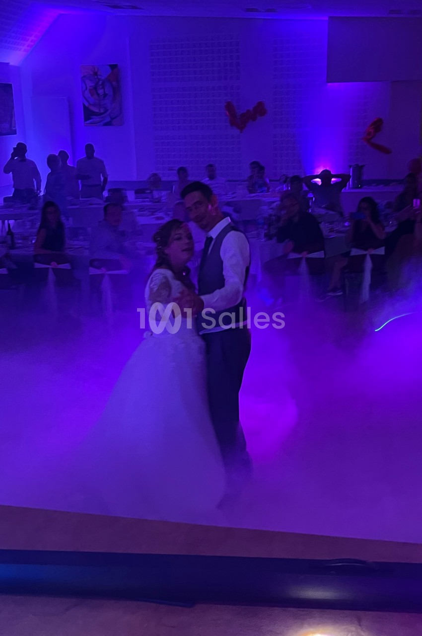 Un couple danse dans une salle éclairée par des lumières violettes, entouré de fumée et d'invités assis.