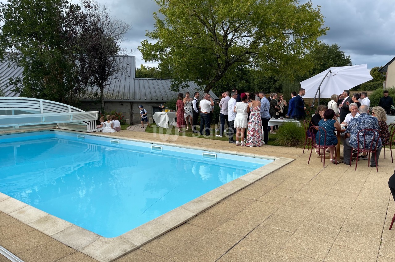 Groupe de personnes réunies près d'une piscine extérieure, avec des arbres et des tables dressées en arrière-plan.