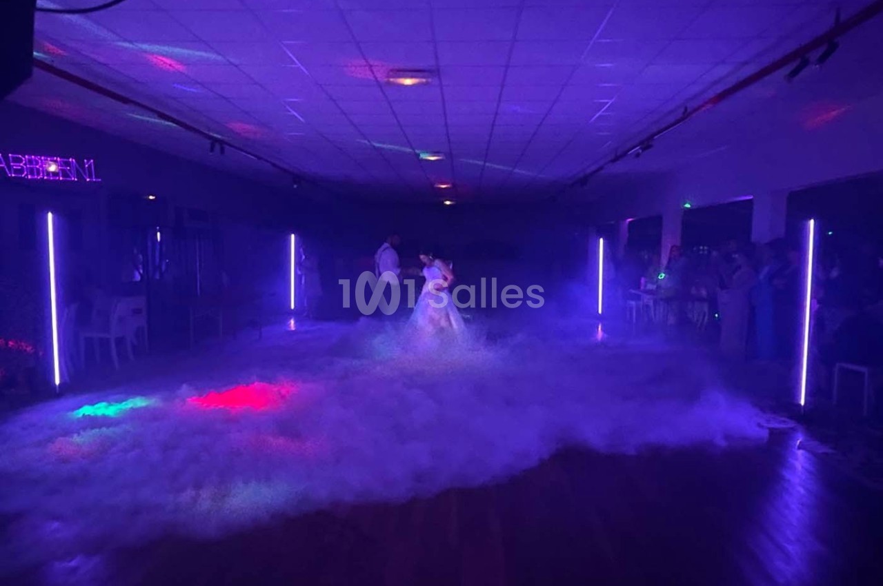 Un couple danse au centre d'une piste enveloppée de fumée, éclairée par des lumières colorées dans une salle sombre.