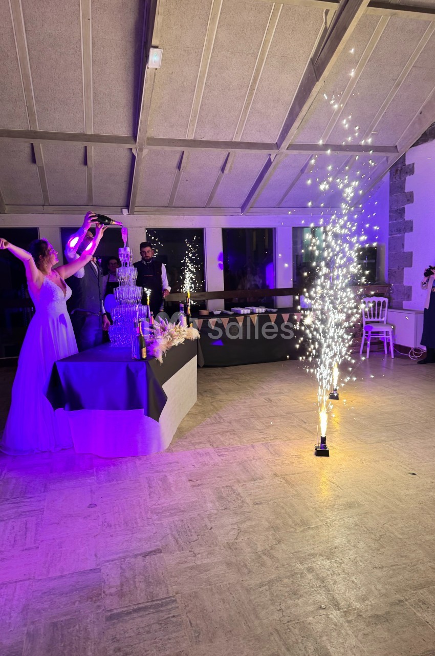 Une femme en robe blanche lève les bras près d'une fontaine de champagne et de feux d'artifice en intérieur.