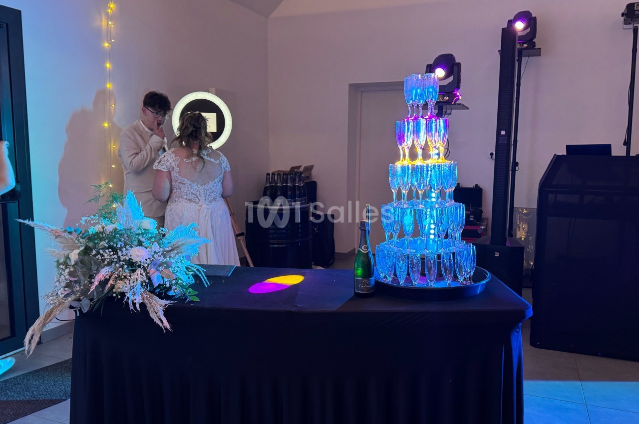 Tour de flûtes à champagne illuminées sur une table noire, décorée de fleurs, dans une salle de réception.