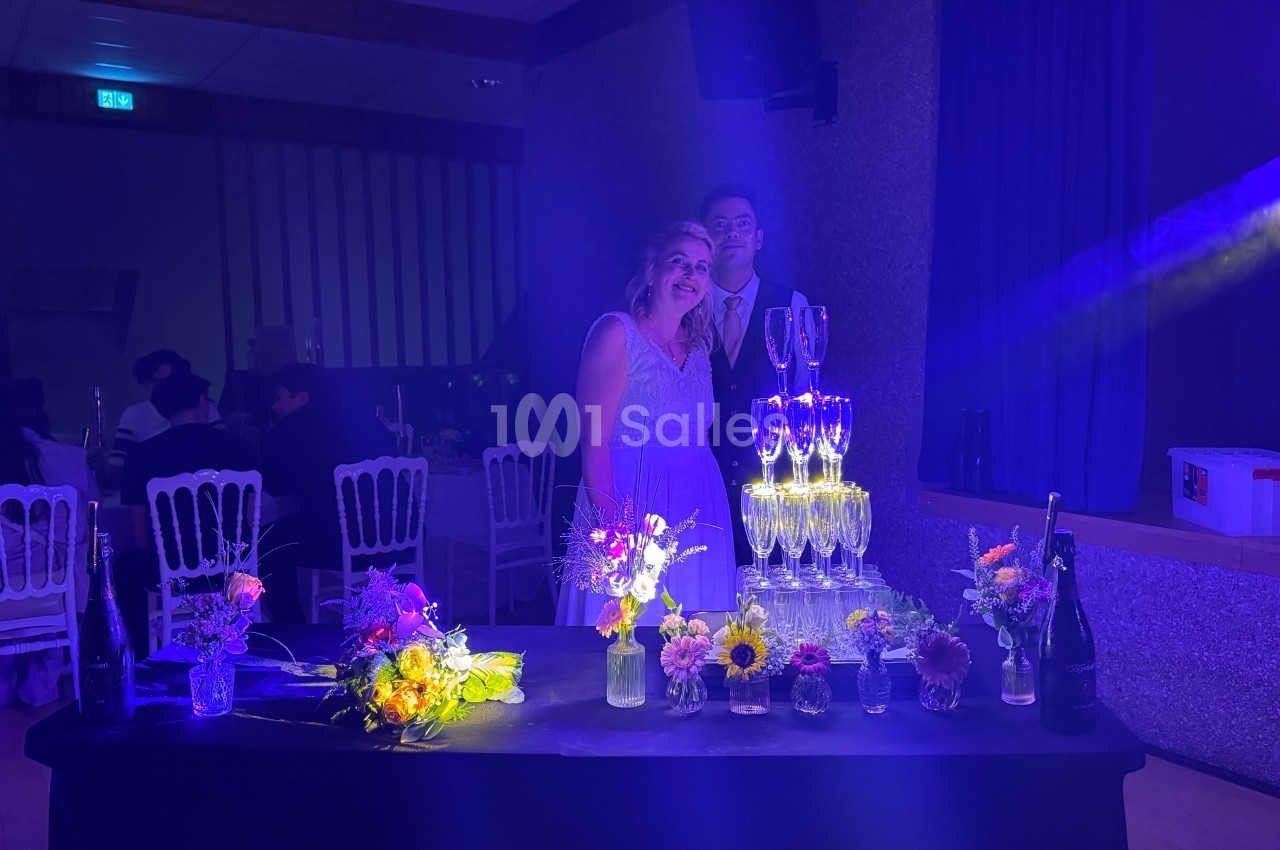 Un couple pose près d'une table décorée de fleurs et d'une pyramide de coupes de champagne dans une salle éclairée.