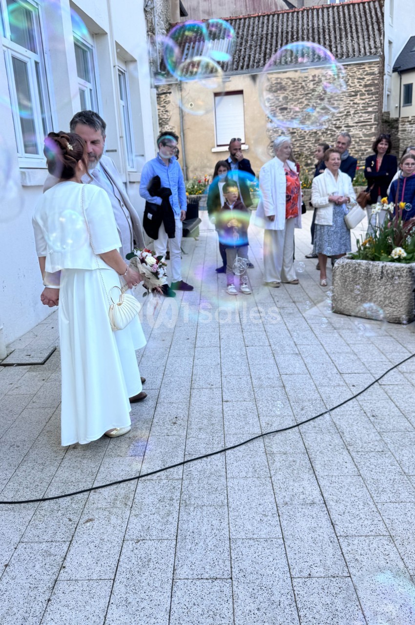 Un couple en tenue de mariage se tient devant un groupe de personnes sur une place pavée, entouré de bulles de savon.