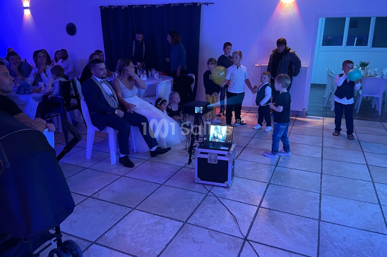 Des invités, dont des enfants, assistent à une projection vidéo dans une salle décorée pour une célébration.