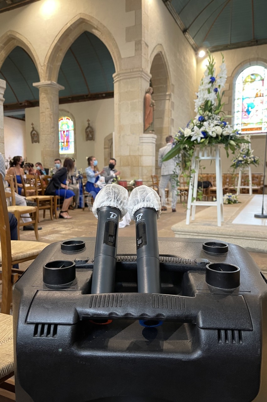 Deux micros sans fil posés sur une enceinte dans une église, avec des invités assis et des décorations florales en arrière…