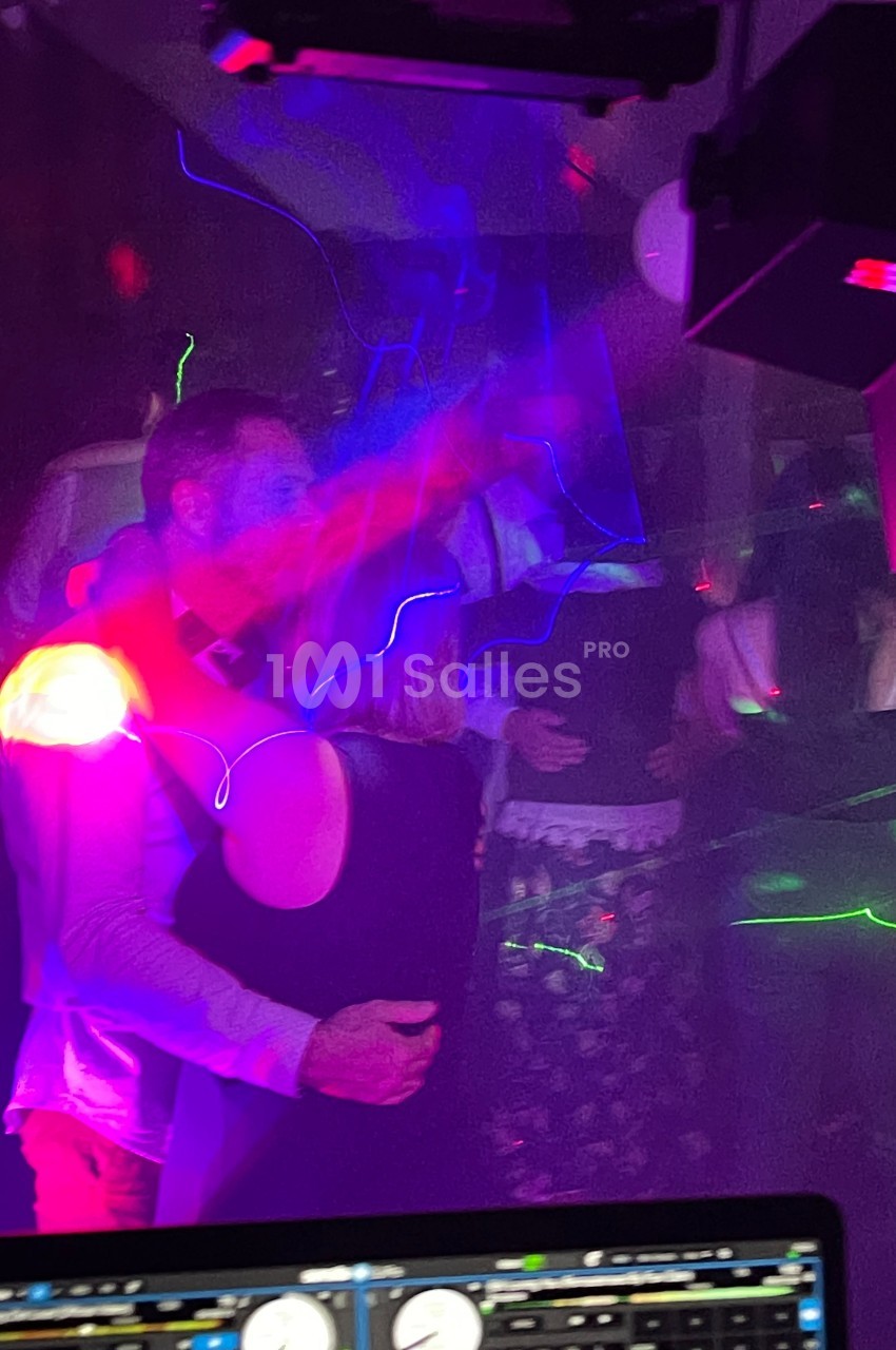 Un couple danse sous des lumières colorées dans une ambiance de fête avec d'autres personnes en arrière-plan.