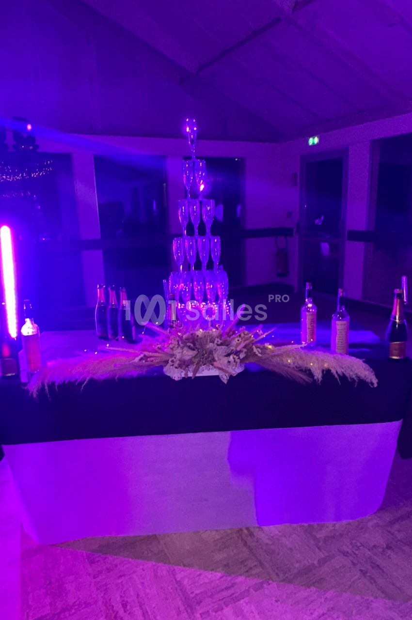 Tour de coupes en verre empilées sur une table décorée, entourée de bouteilles, éclairée par une lumière violette.