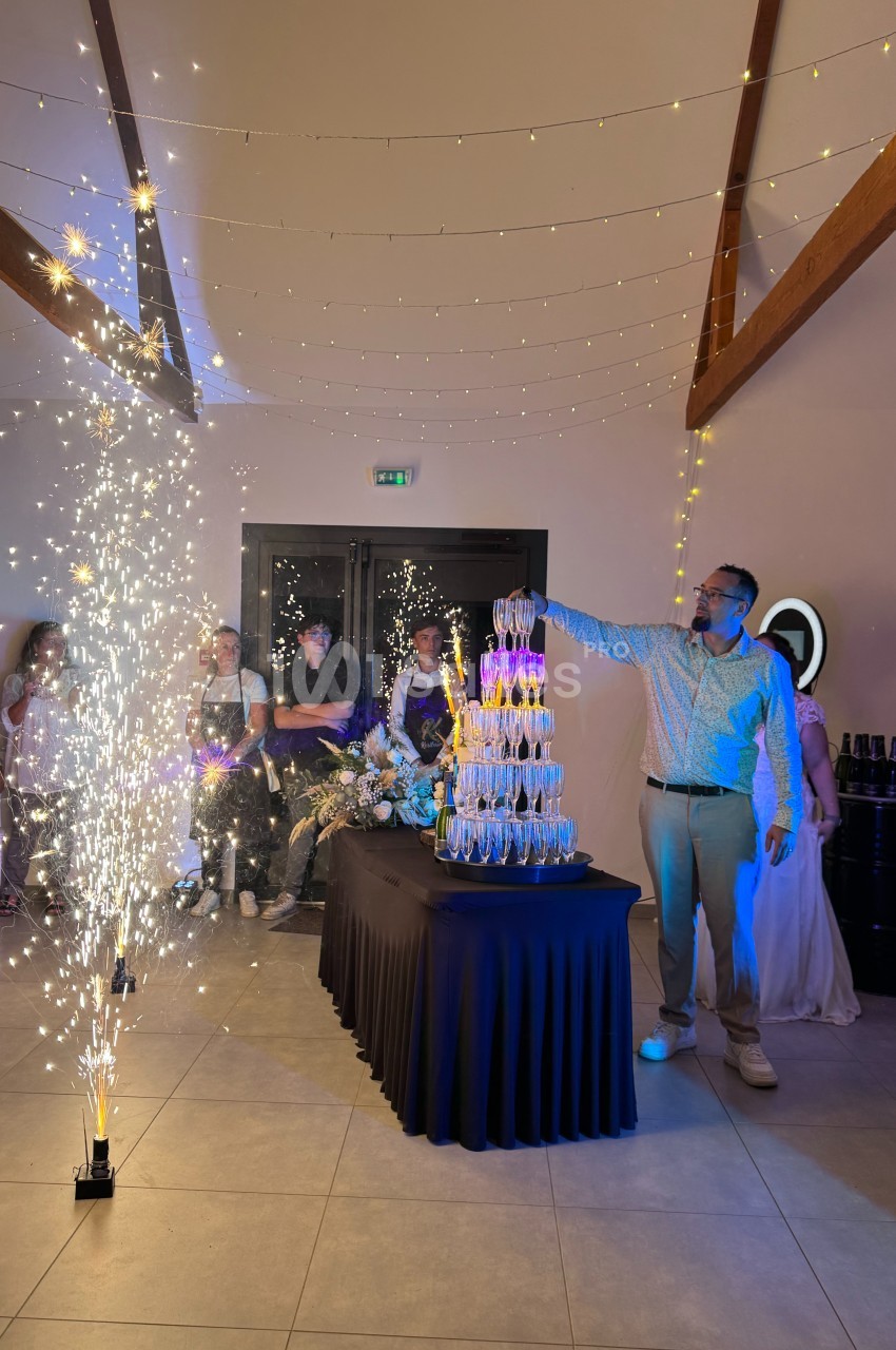 Un homme verse du champagne sur une pyramide de verres, entouré de convives et de décorations lumineuses.