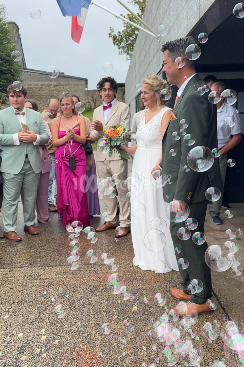 Un couple de mariés entouré d'invités, sous des bulles de savon, devant un bâtiment avec un drapeau français.