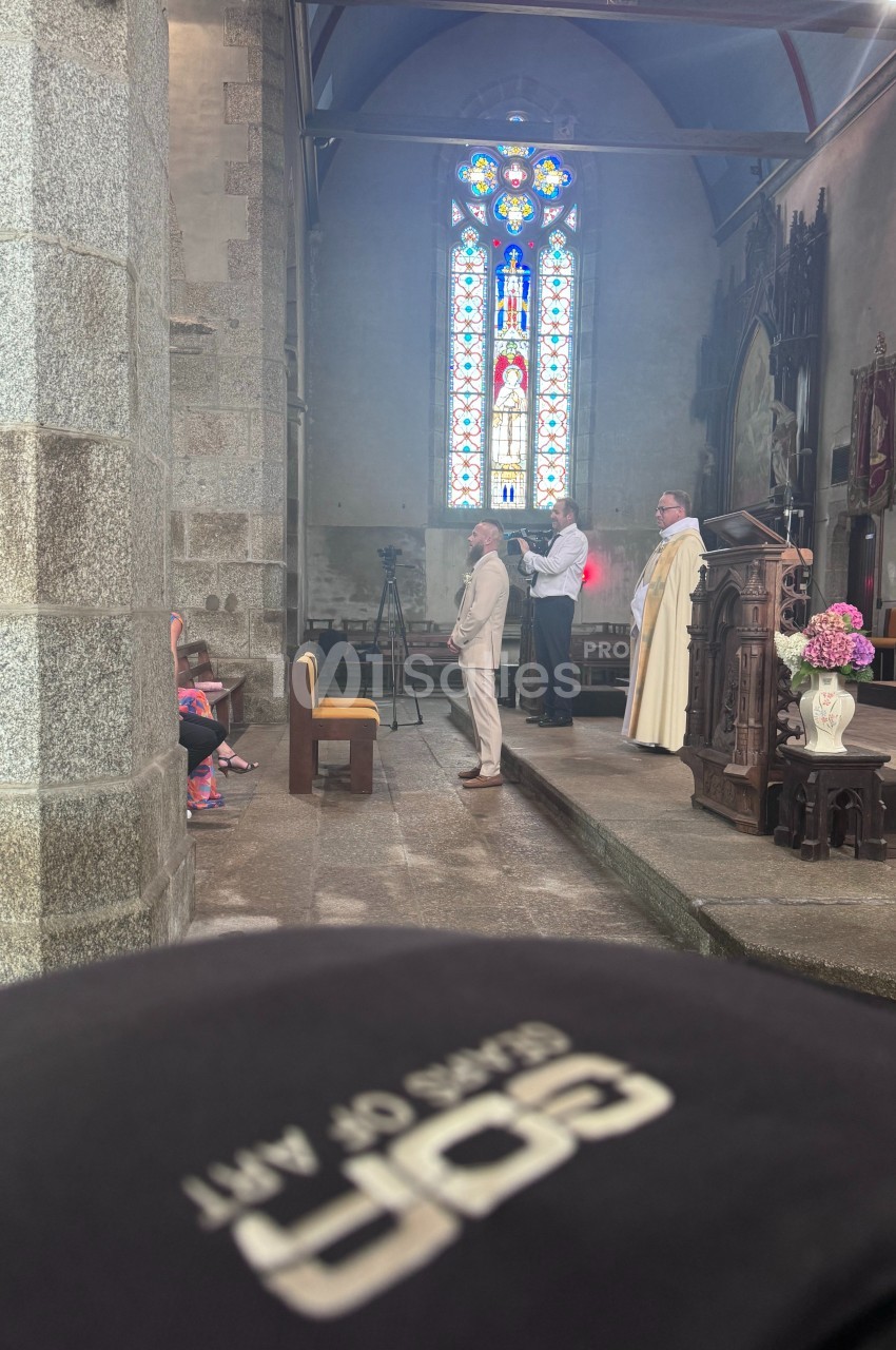 Un homme en costume est baptisé par un prêtre dans une église avec un vitrail coloré en arrière-plan.