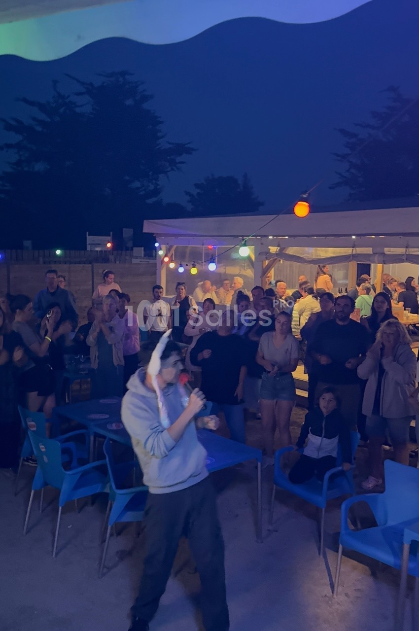 Un homme chante au micro devant un public assis et debout, éclairé par des guirlandes lumineuses en soirée.
