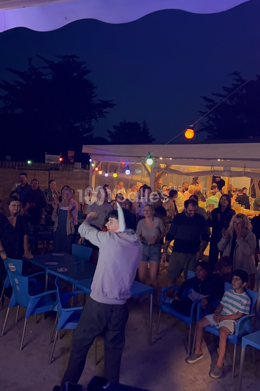 Un homme avec un micro anime une soirée en plein air devant un public assis et debout sous des guirlandes lumineuses.