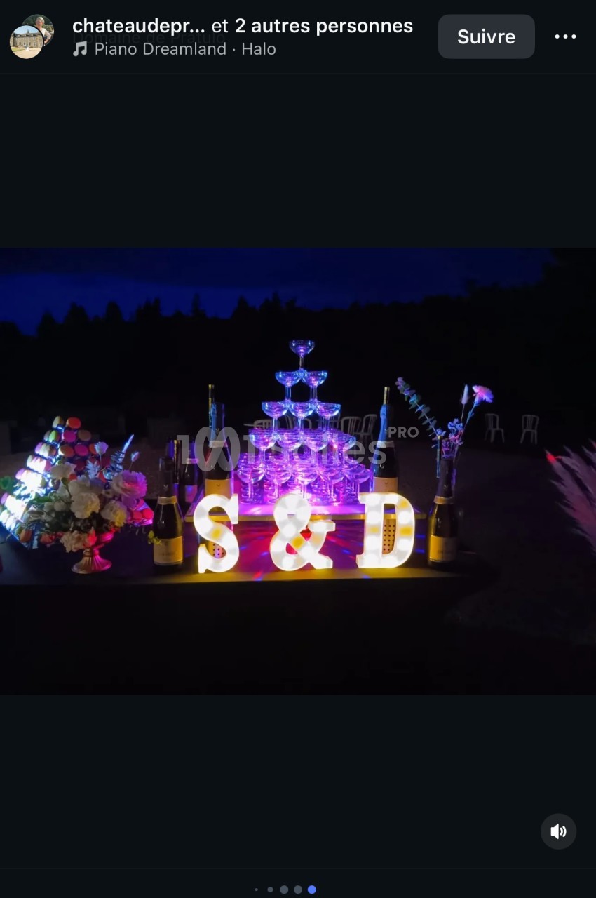 Tour de coupes de champagne illuminée entourée de bouteilles, fleurs et lettres décoratives ’S & D’ dans un décor nocturne.