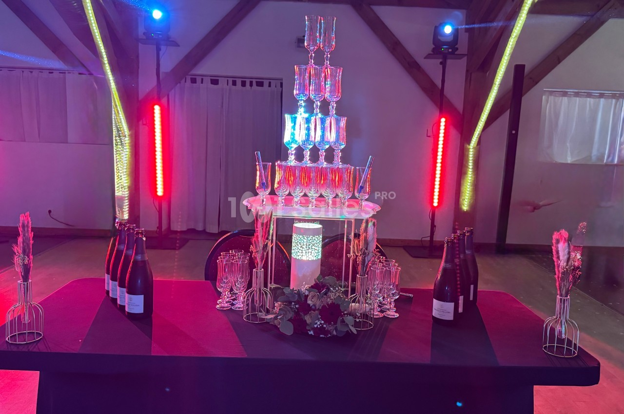 Tour de coupes en verre empilées sur une table décorée, entourée de bouteilles et éclairée par des lumières colorées.