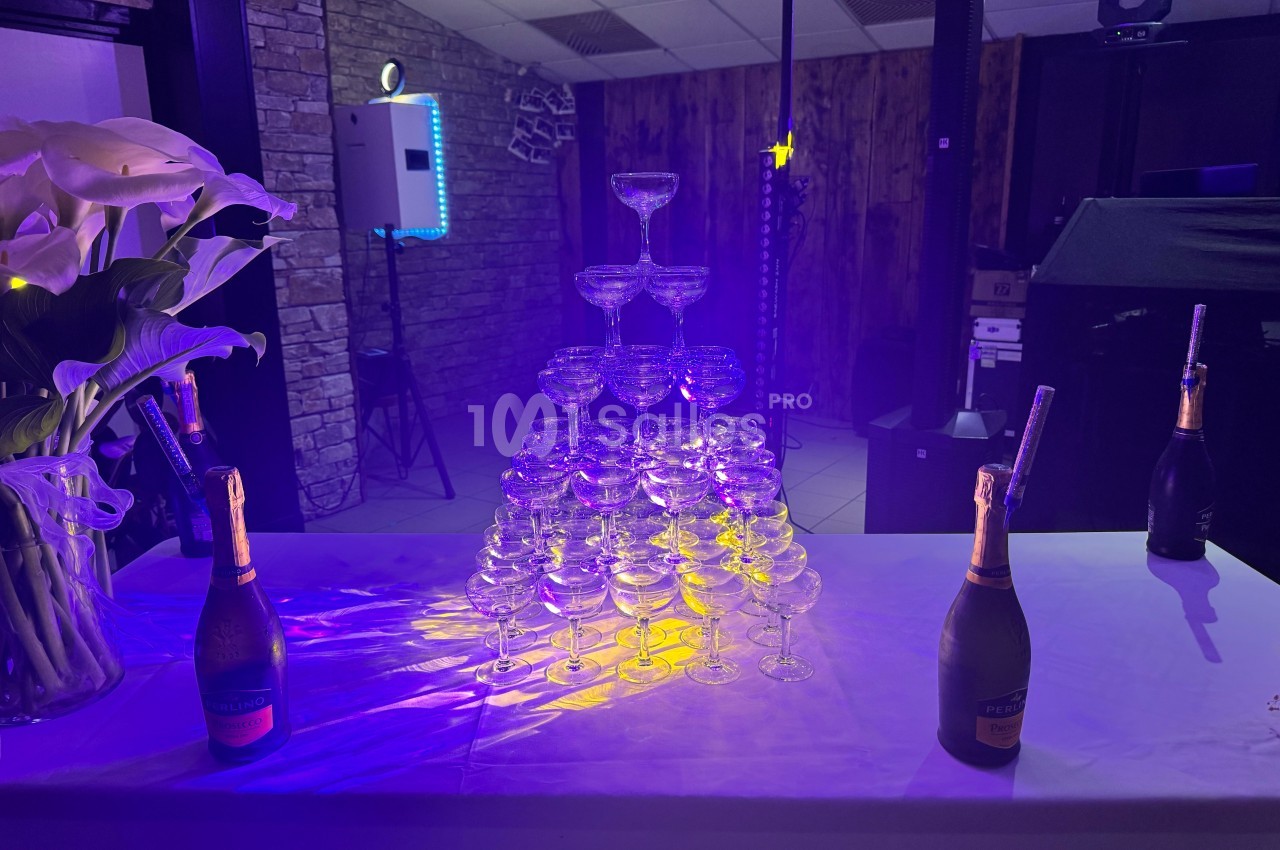 Pyramide de coupes en verre éclairée, entourée de bouteilles de champagne sur une table blanche dans une salle décorée.