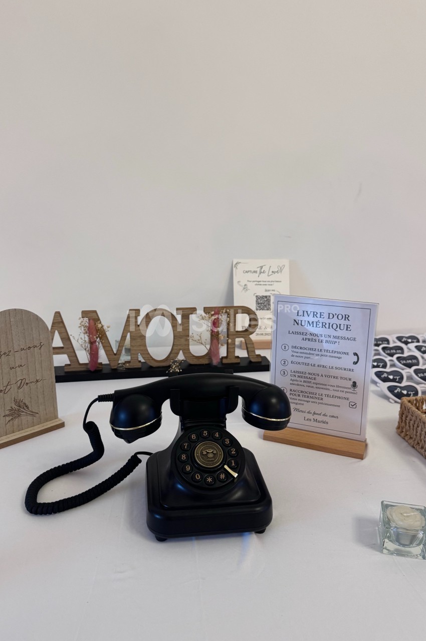 Téléphone rétro noir sur une table décorée avec des objets, dont un panneau ’AMOUR’ et un livre d'or numérique.