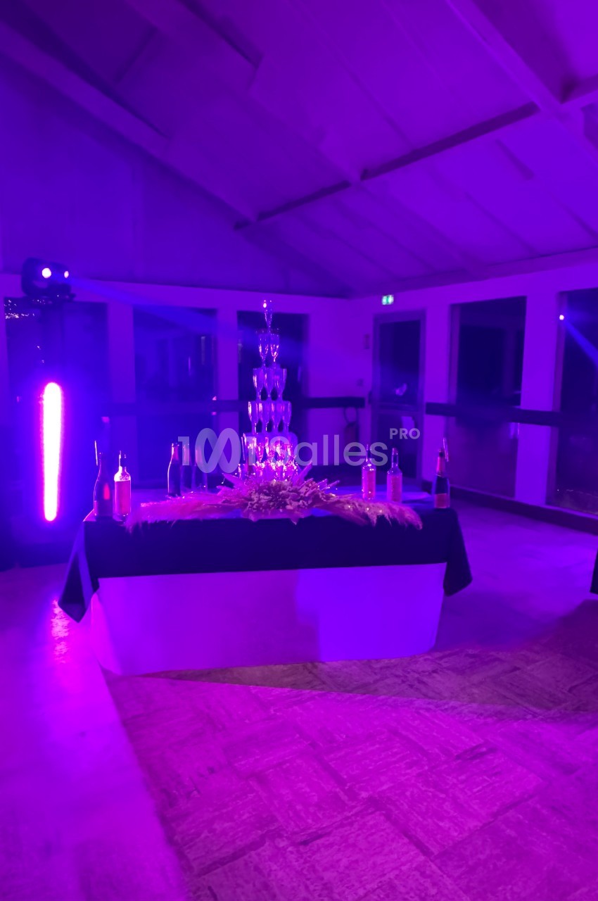 Tour de coupes en verre empilées sur une table décorée, entourée de bouteilles, éclairée par une lumière violette.