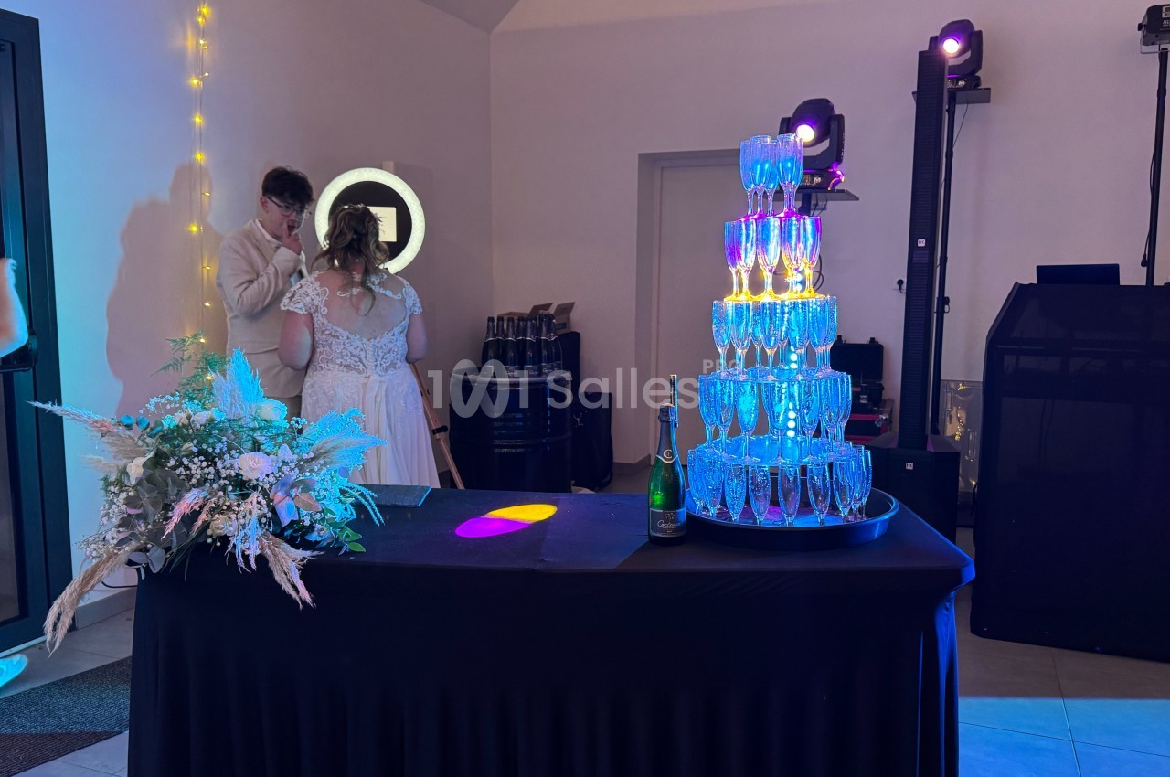 Tour de flûtes à champagne illuminées sur une table noire, décorée de fleurs, dans une salle de réception.