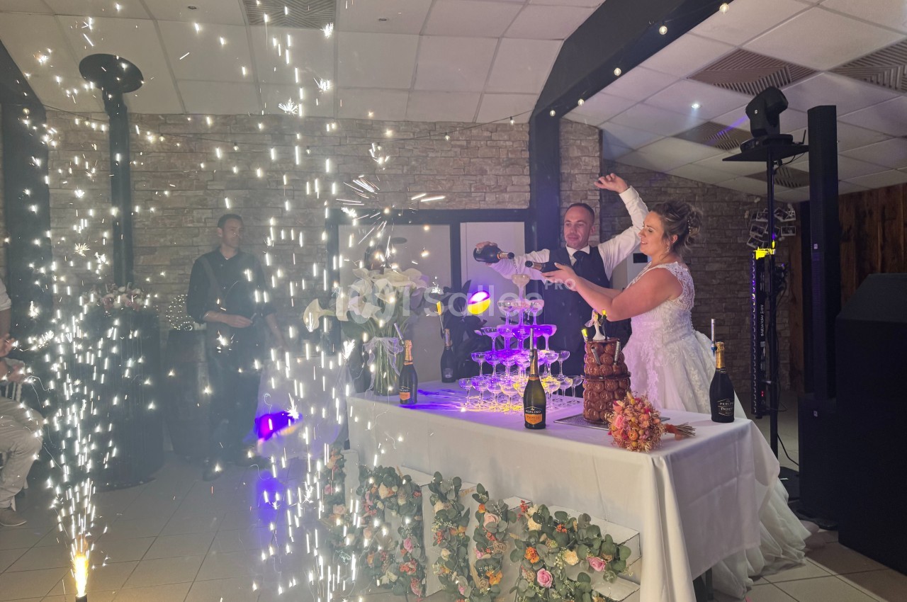 Un couple de mariés verse du champagne dans une pyramide de verres, entouré de décorations et d'effets lumineux.