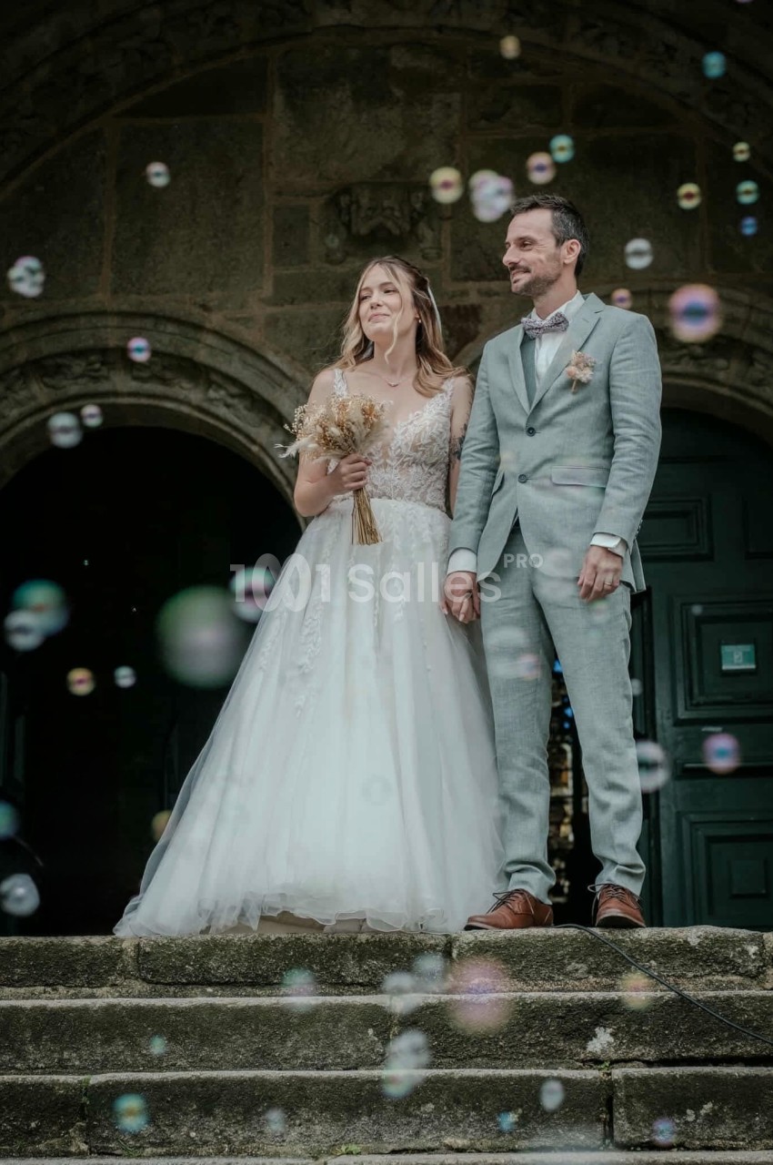 Un couple en tenue de mariage se tient sur les marches d'un bâtiment ancien, entouré de bulles de savon.