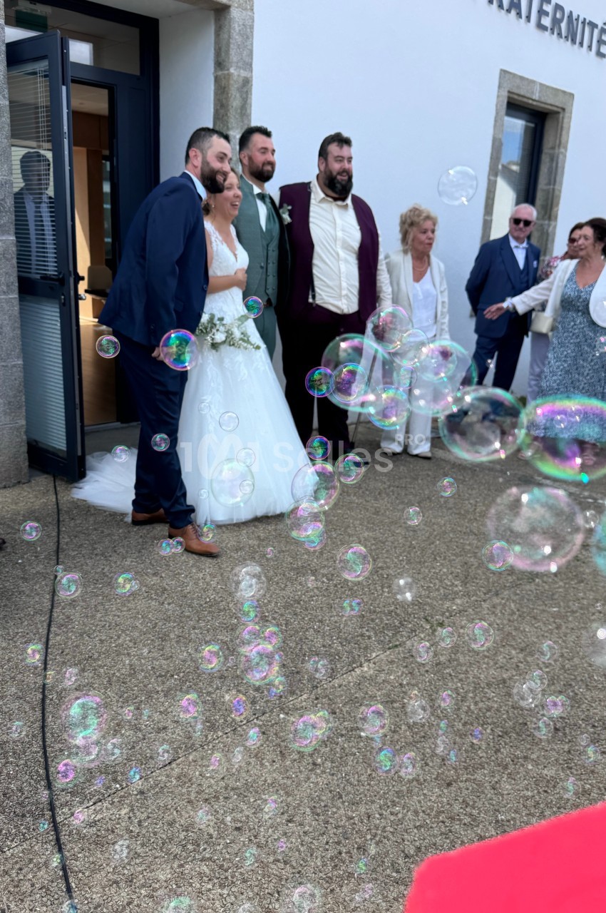 Un couple de mariés pose avec des invités devant un bâtiment, entourés de bulles de savon.