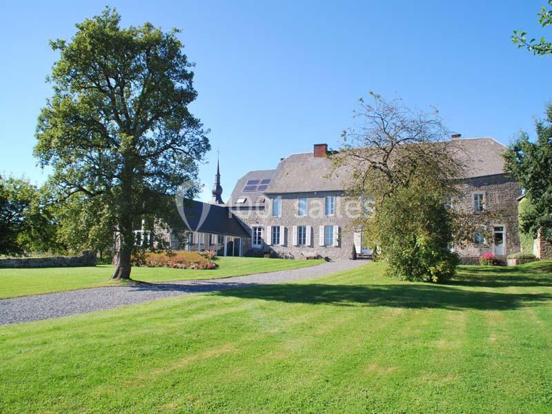 Grande maison en pierre avec volets bleus, entourée d'arbres et d'une pelouse, sous un ciel dégagé.