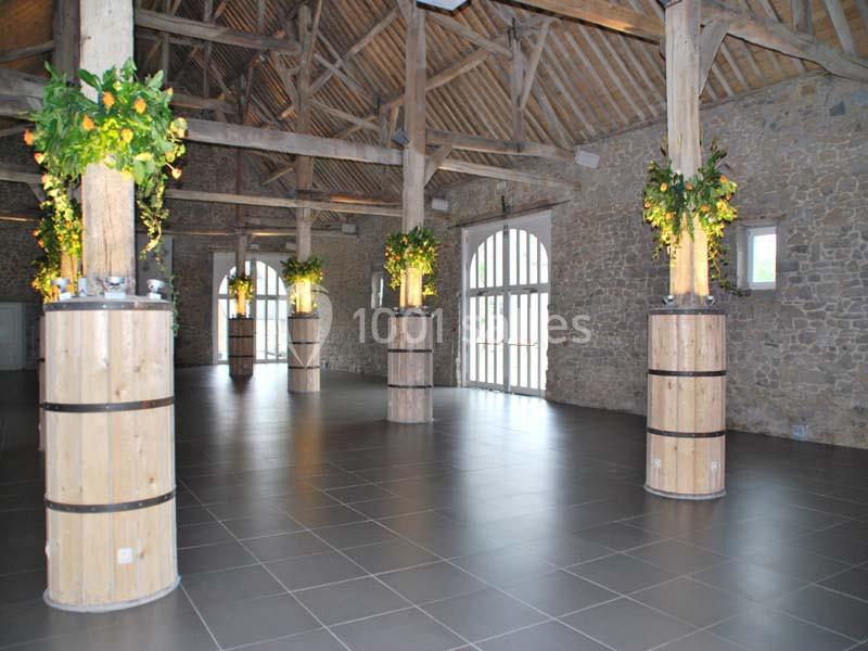 Salle spacieuse avec poutres apparentes, piliers en bois décorés de plantes et grandes fenêtres laissant entrer la lumière.