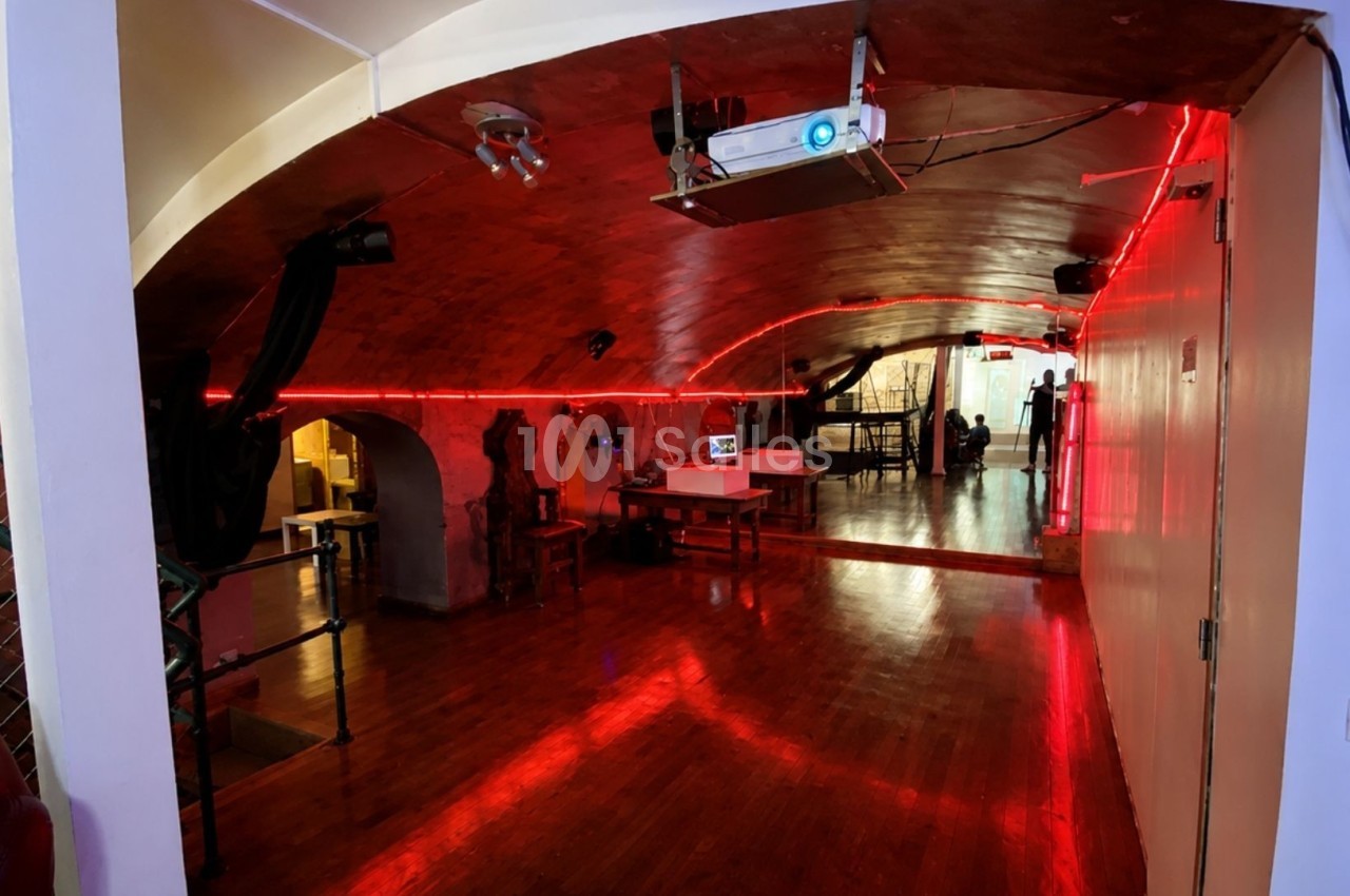 Salle voûtée avec éclairage rouge, parquet en bois, projecteur au plafond et espace aménagé pour des activités.