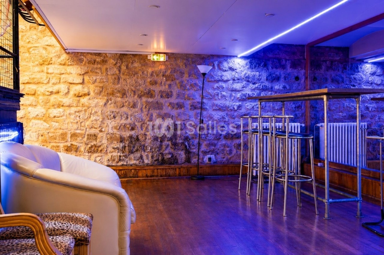Salle avec murs en pierre, éclairage bleu, tables hautes en métal et fauteuils, sol en parquet.