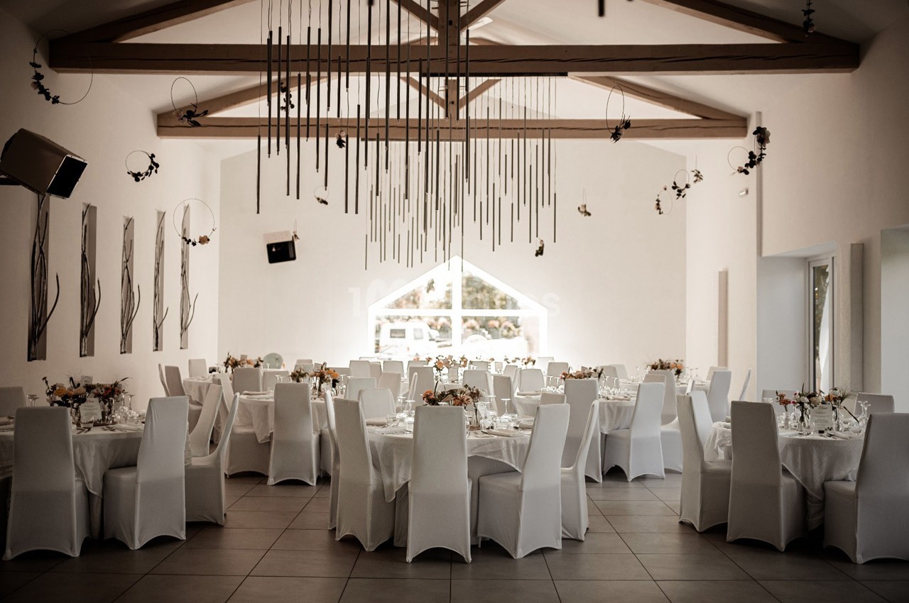 Salle de réception lumineuse avec tables rondes dressées, chaises blanches et décorations suspendues au plafond.