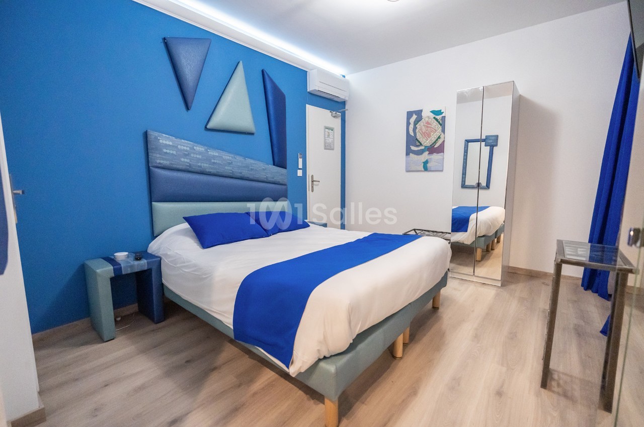 Chambre moderne avec lit double, décor bleu et blanc, sol en bois clair, miroir et éléments géométriques au mur.