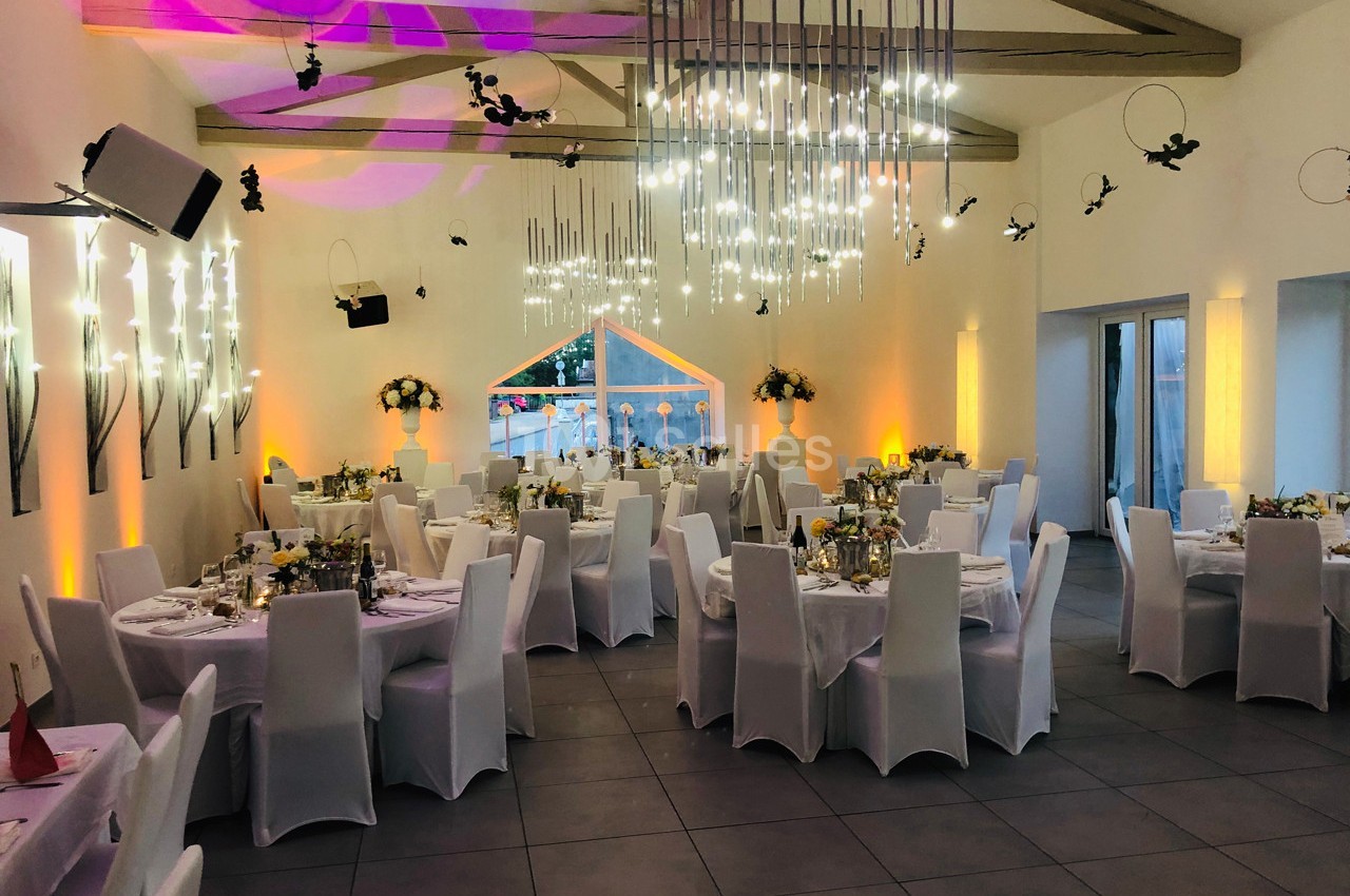 Salle de réception décorée avec des tables rondes, nappes blanches, chaises assorties et éclairage moderne.