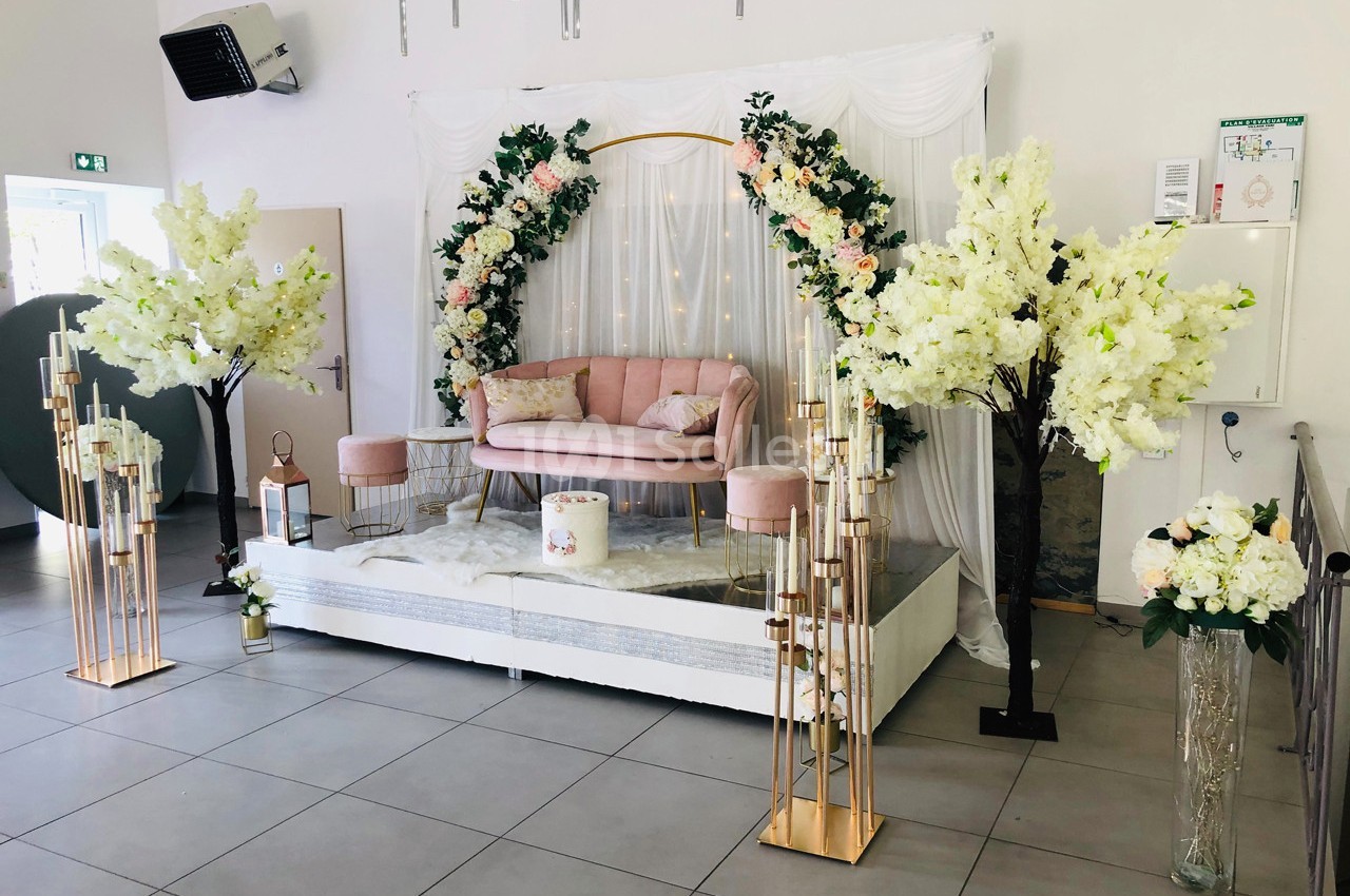 Décoration de salle avec arche florale, canapé rose, fleurs blanches et dorures sur une estrade.