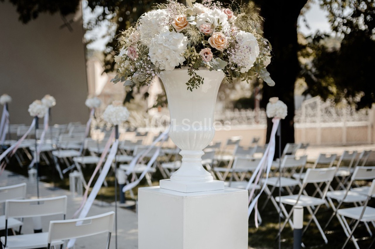 Arrangement floral dans un vase blanc sur un piédestal, décorant une allée bordée de chaises pour une cérémonie en extérieur.